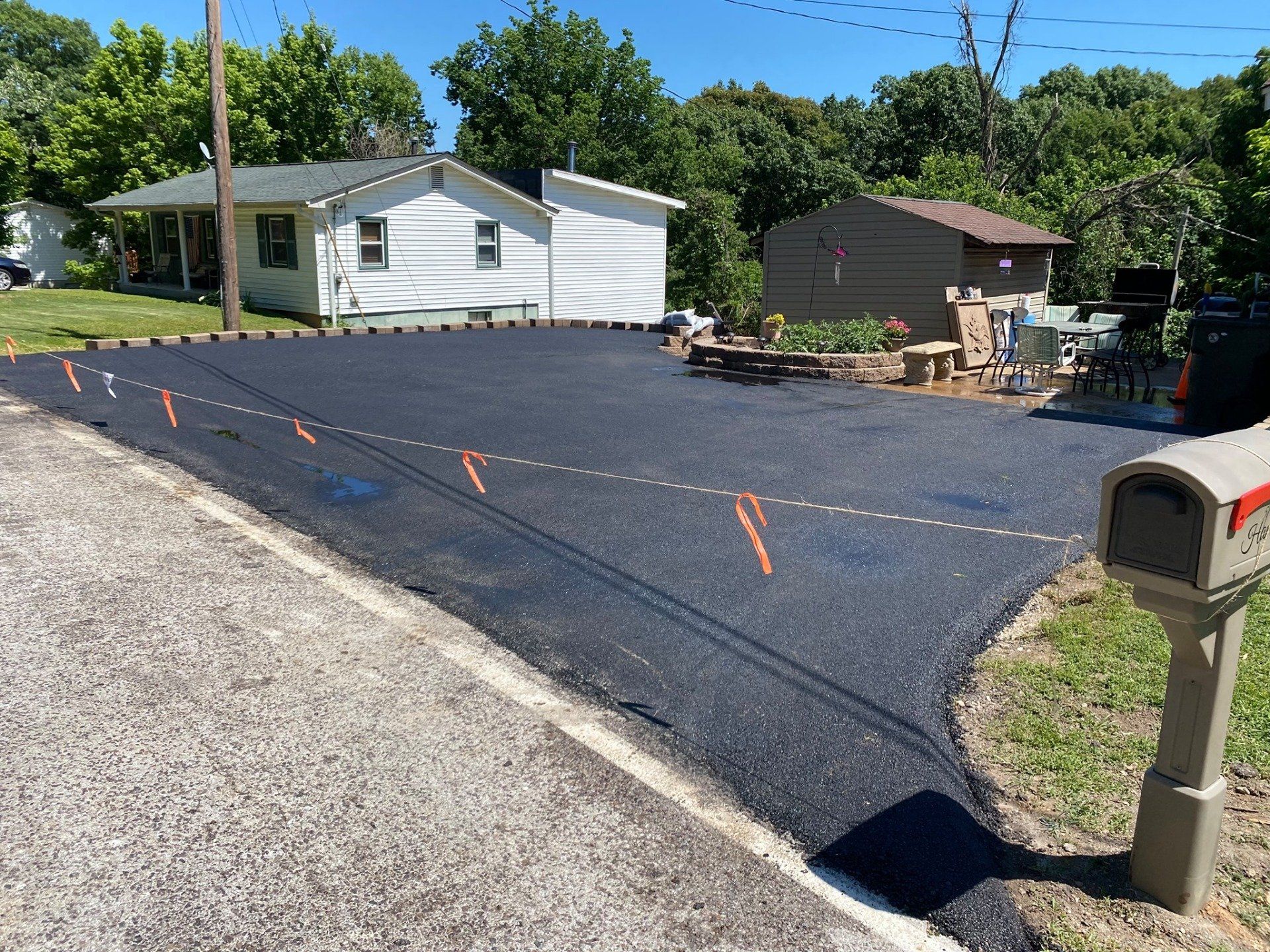Freshly paved black driveway beside a white house, with a mailbox and trees nearby