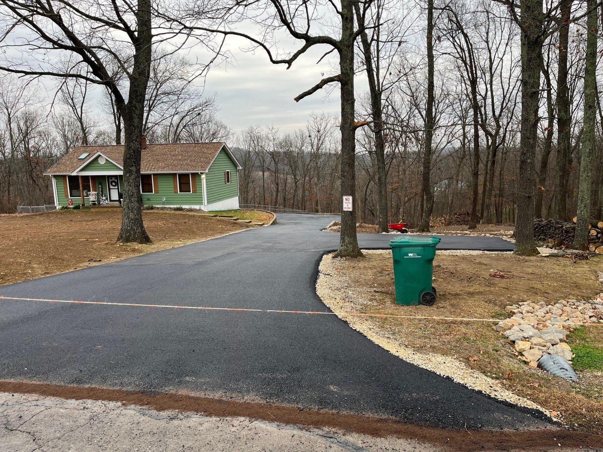 Paved driveway leading to a green house in a wooded yard, with a trash bin and bare trees.