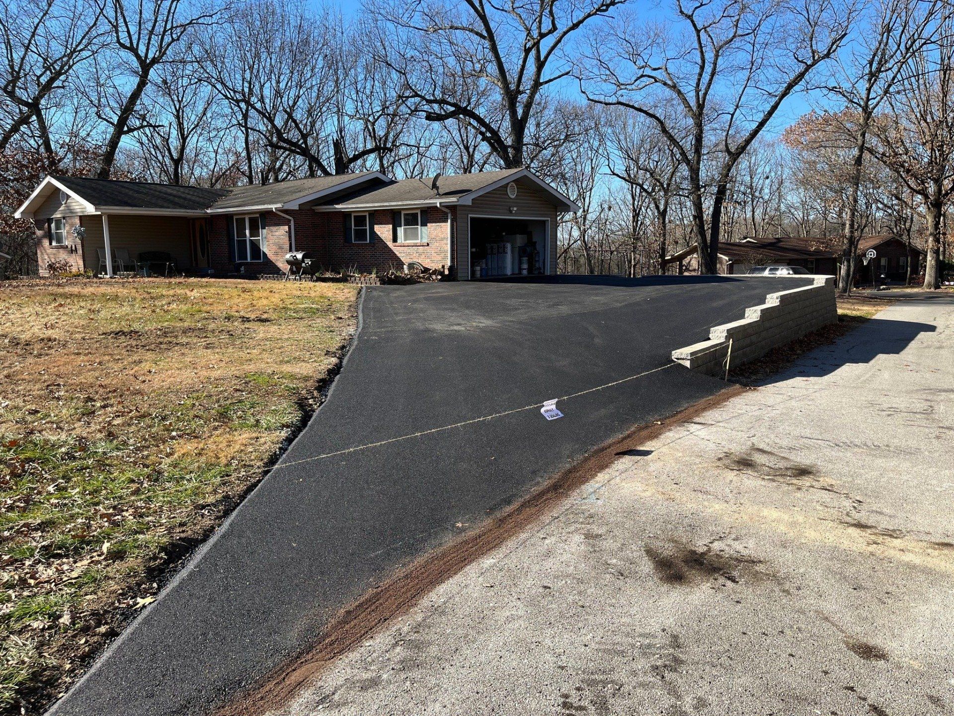 Steep black asphalt driveway beside a house, bordered by a concrete retaining wall and leafless trees.