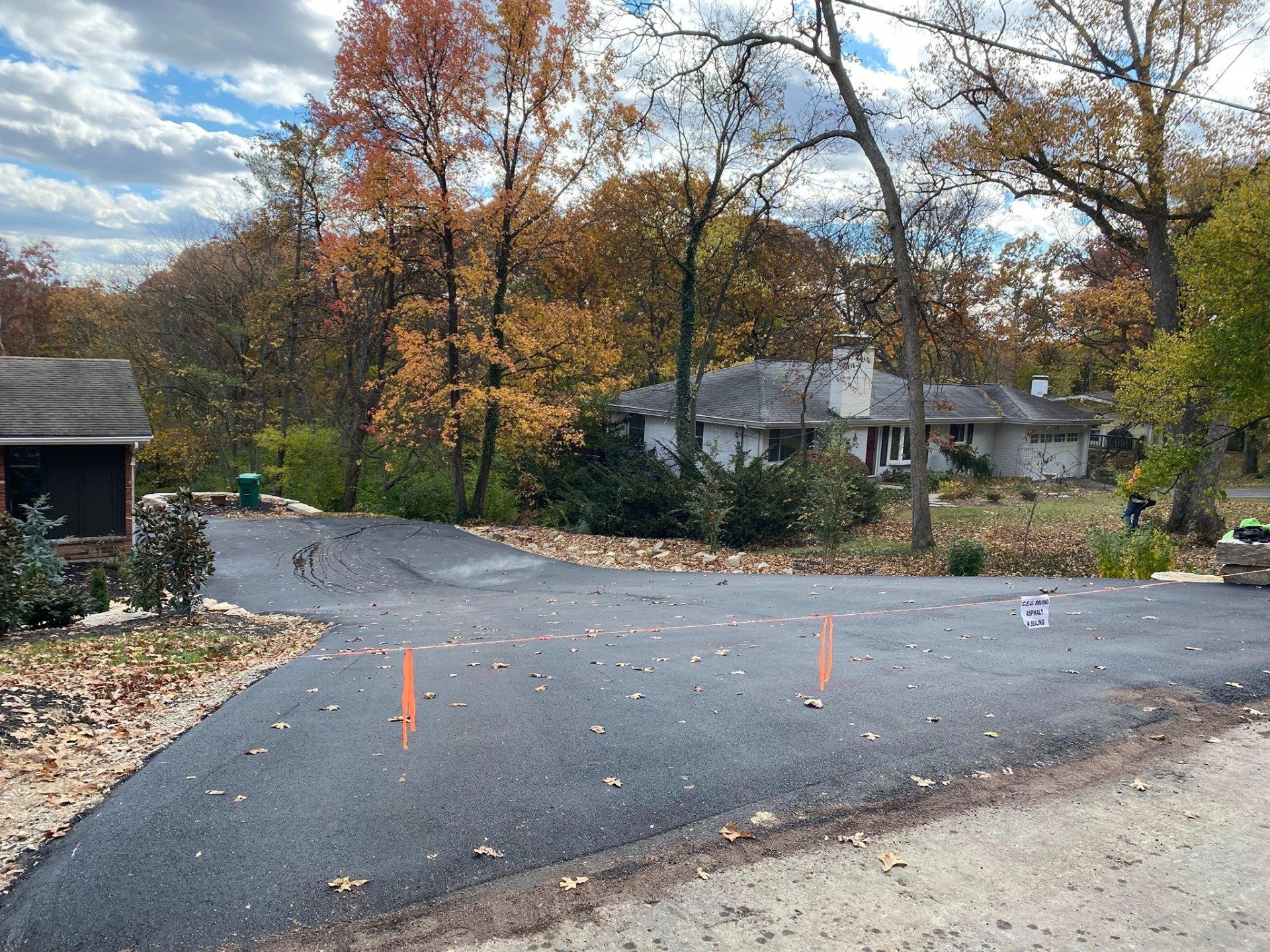 Curved asphalt driveway under construction, marked with orange cones, beside a house and autumn trees.