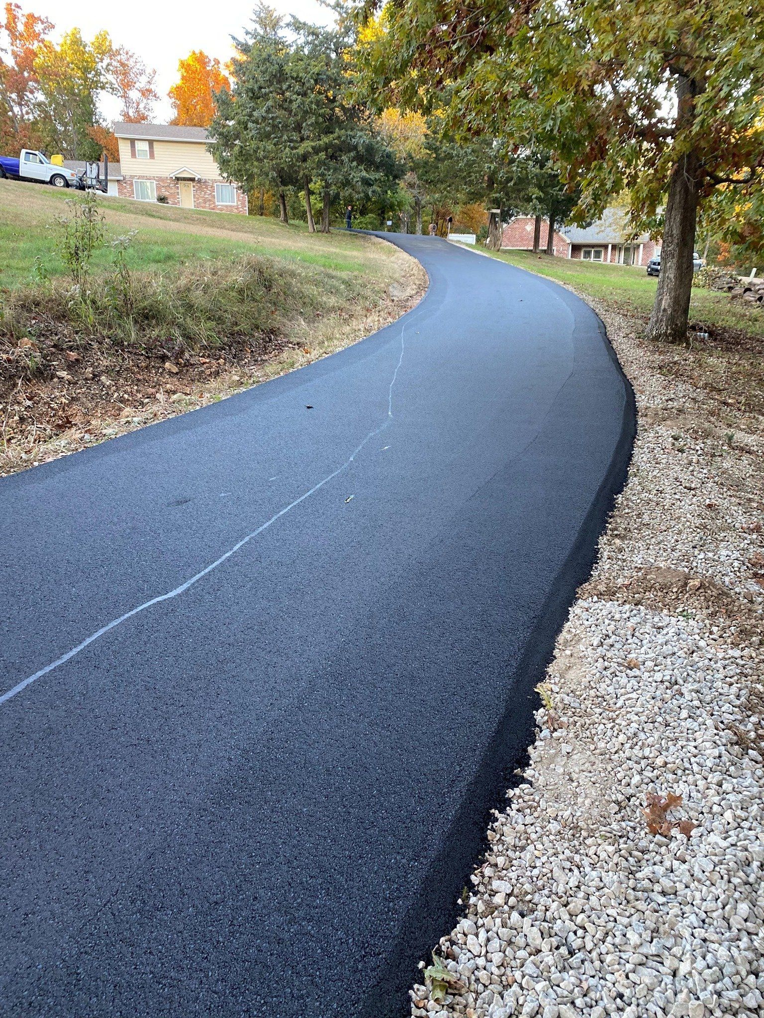 Curved black asphalt path through a grassy park with trees and fallen leaves in autumn.