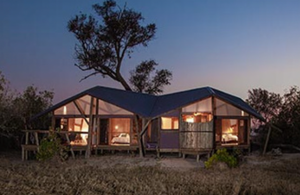 Safari lodge at dusk, showcasing lit interiors and dark blue roof.  The wooden structure is set in a grassy field with trees.