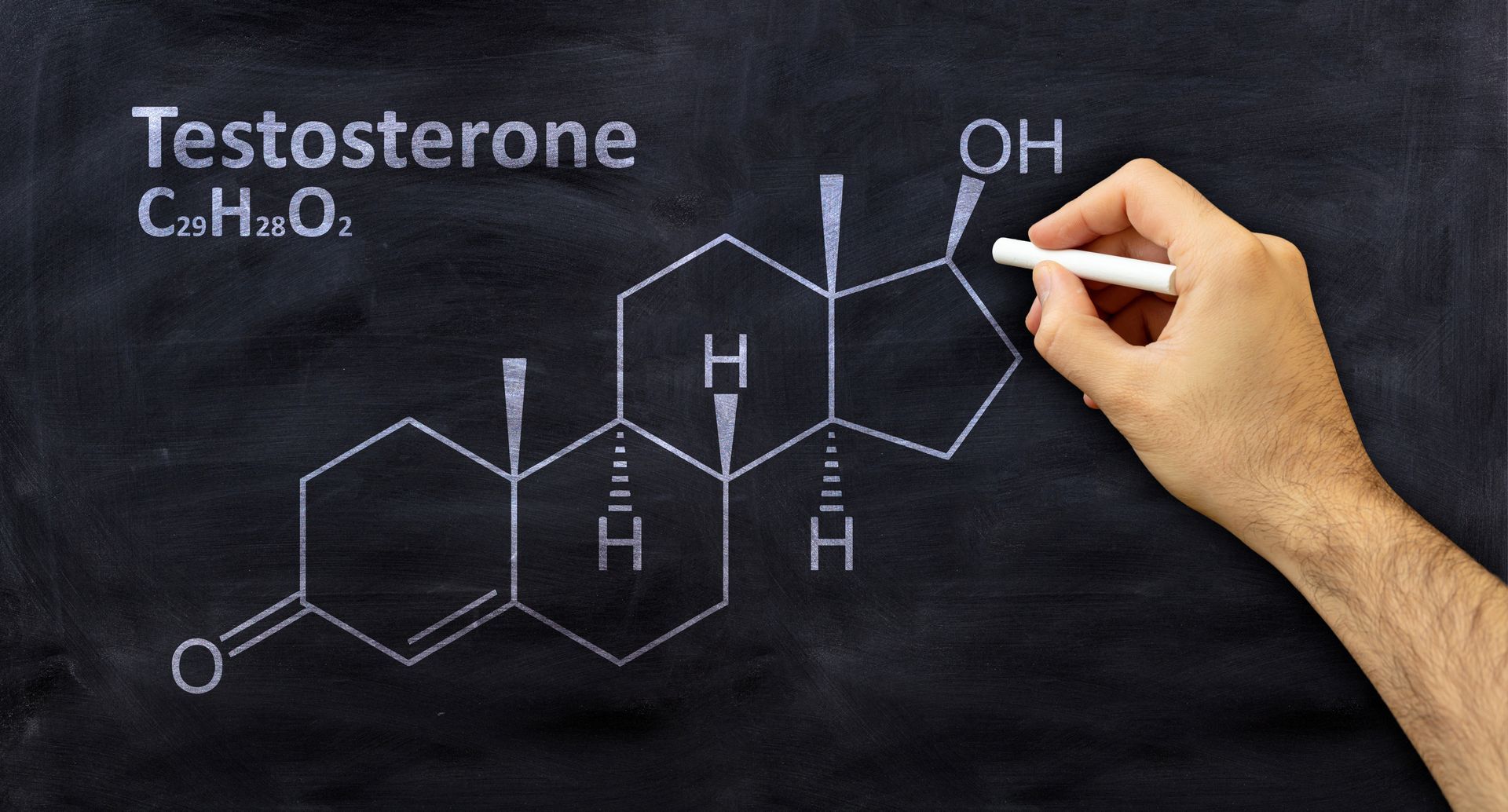 A hand is drawing the chemical structure of testosterone on a blackboard