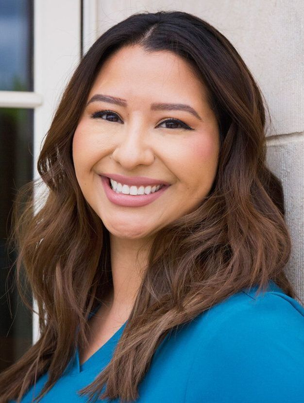 A woman in a blue shirt is smiling and leaning against a wall.