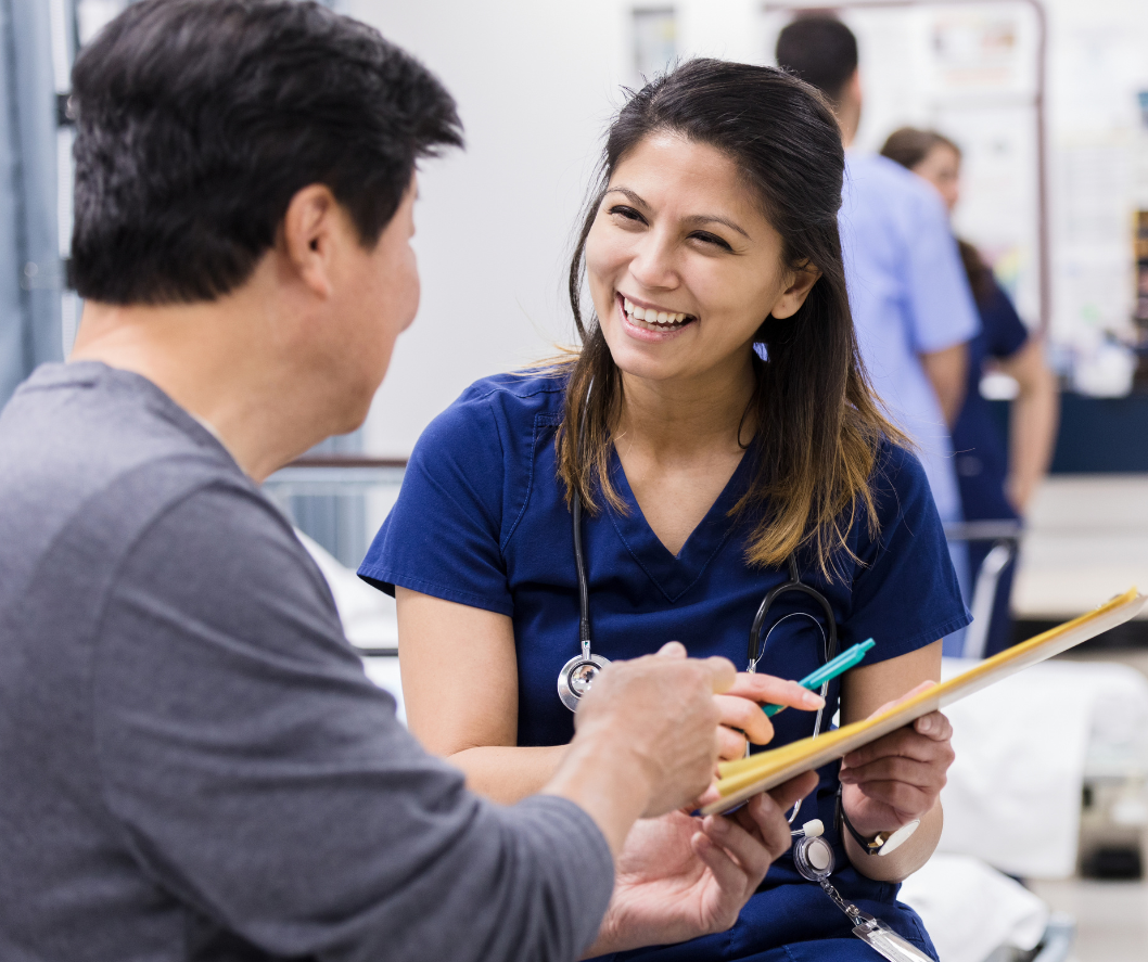 A nurse is talking to a patient in a hospital room while holding a clipboard.