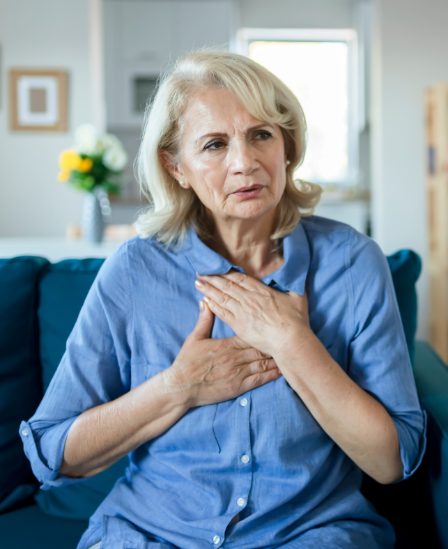 An elderly woman is sitting on a couch with her hands on her chest.