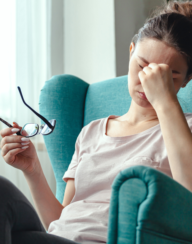 A woman is sitting in a chair holding her glasses and rubbing her forehead.