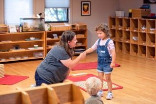 Teacher kneeling to guide a child in a classroom with cubbies and rugs