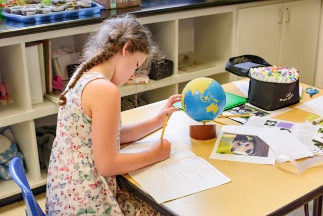 Student writing at a desk with a globe, papers, and art supplies in a classroom