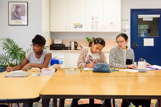 Three people working at a table in a bright classroom, writing and studying with books and notebooks.