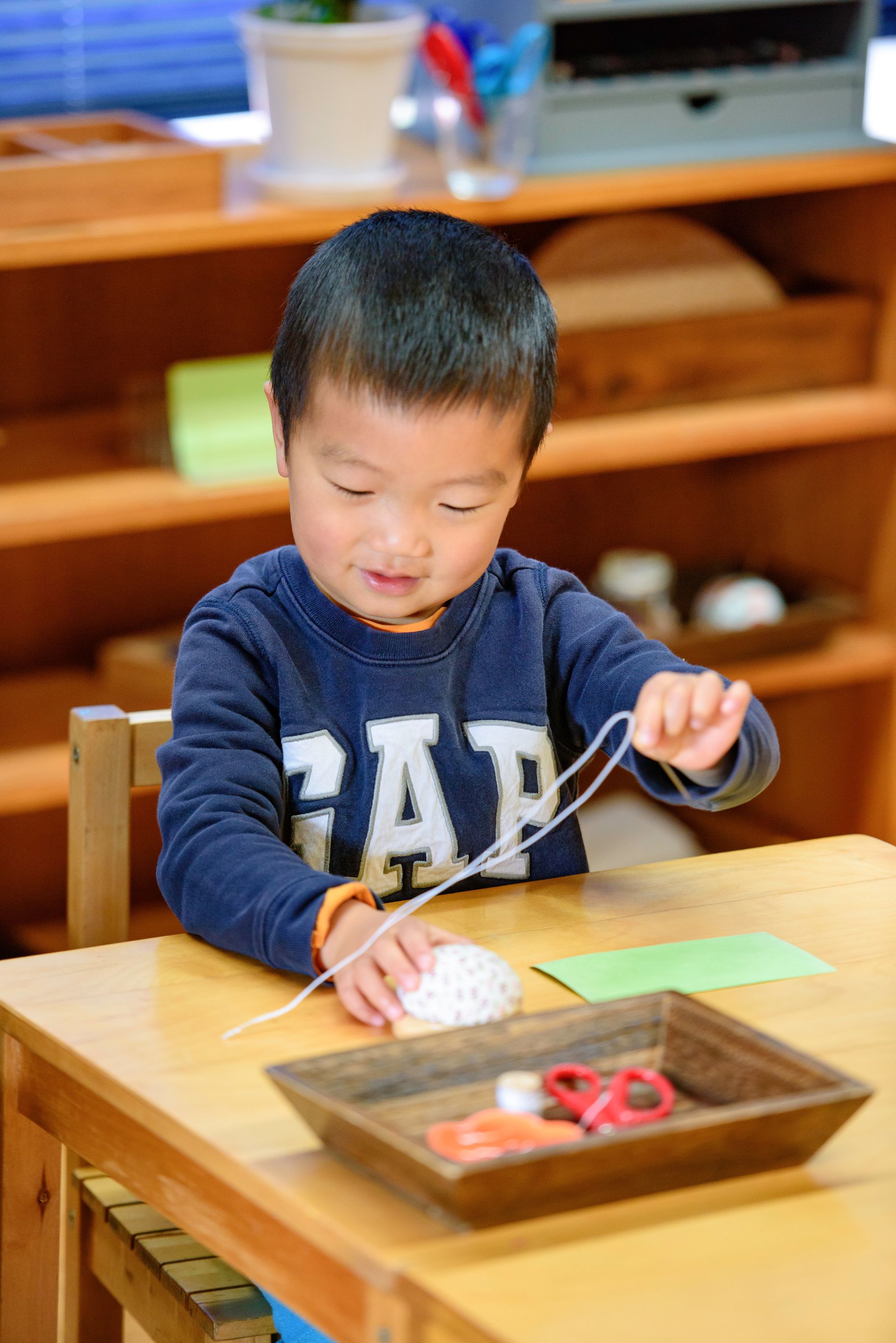 Child threading a string through a bead at a wooden table in a classroom.