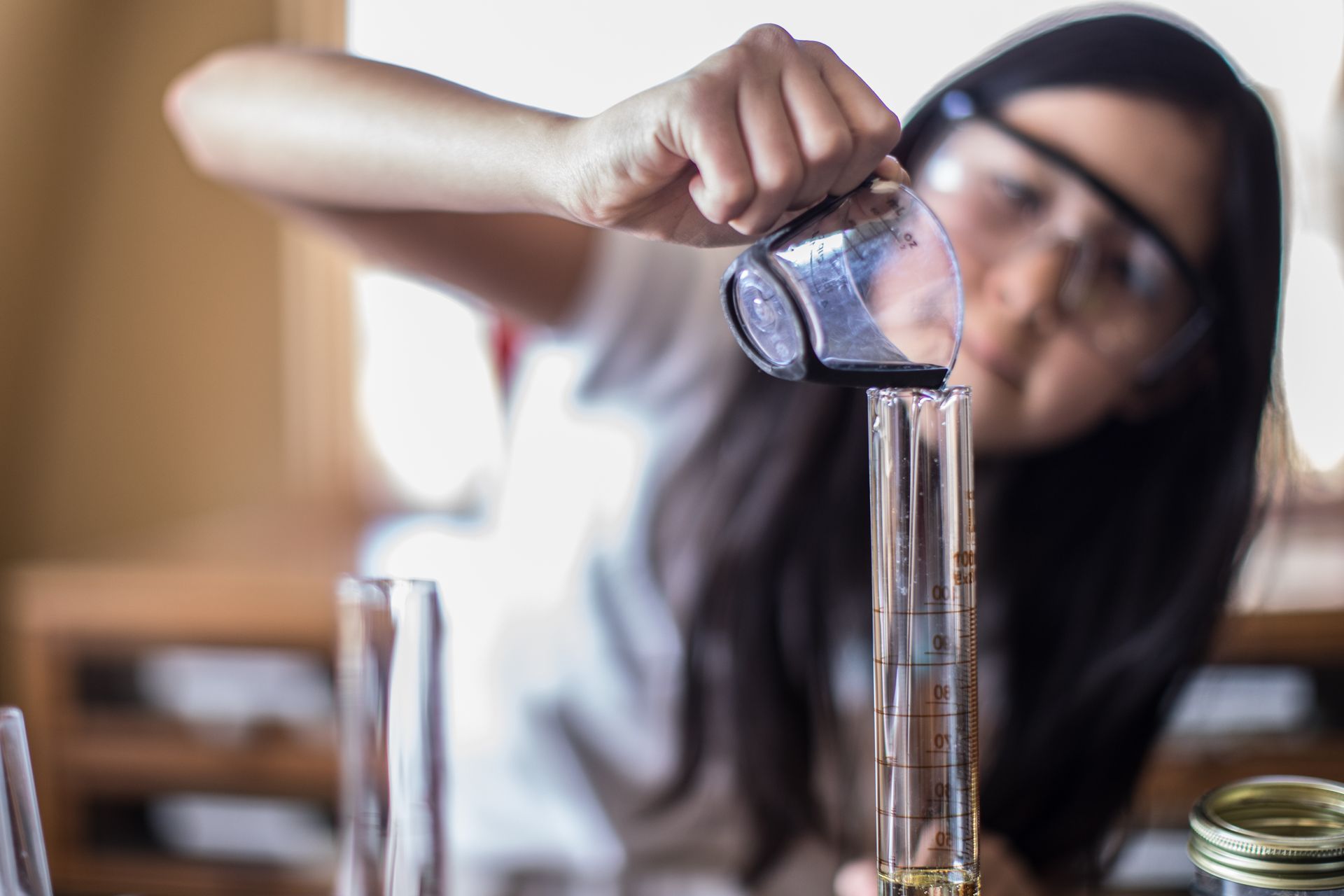 Child pouring clear liquid from a glass into a tall container on a table, indoors