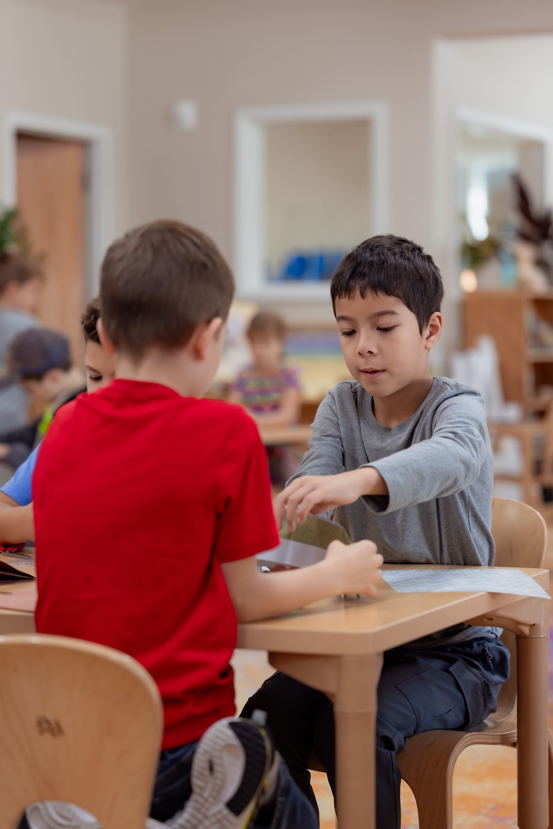 Two children sit at a classroom table, reaching toward a shared activity with paper and small objects.