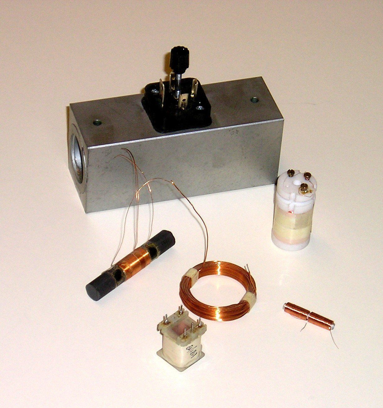 Varie bobine di rame e componenti elettrici, tra cui un solenoide e un blocco metallico rettangolare con un connettore.