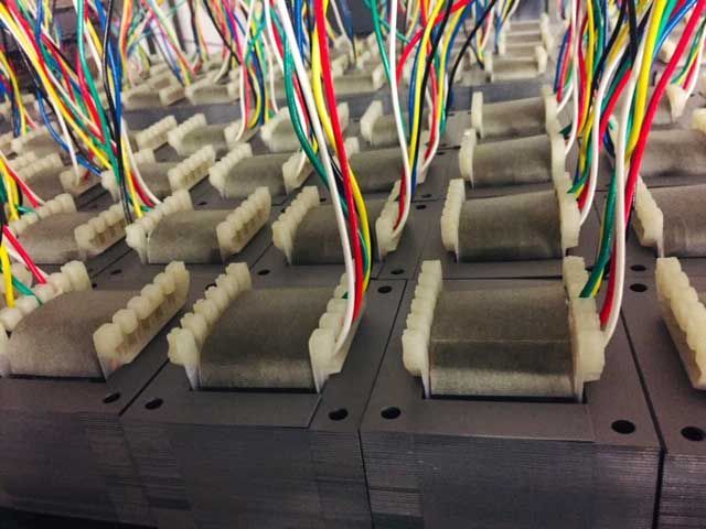 File di componenti elettronici rettangolari grigi con fili colorati attaccati, impilati su piastre metalliche.