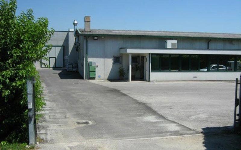 Edificio industriale grigio con vialetto asfaltato e verde, sotto un cielo limpido.
