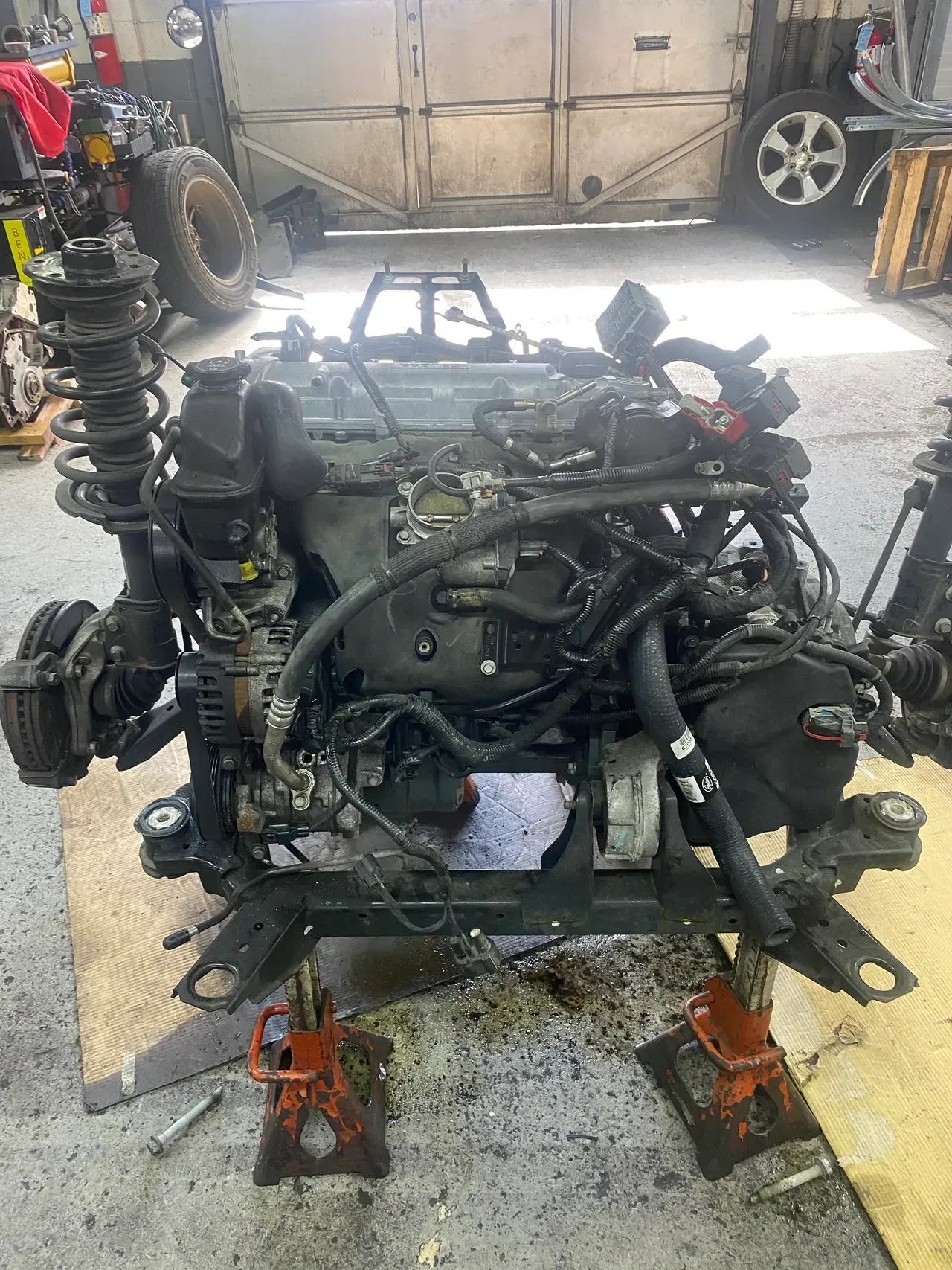 Car engine mounted on a stand in a garage, with tools and a vehicle in the background.
