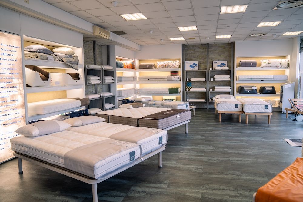 Showroom di materassi con vari letti esposti su piattaforme, luci soffuse e scaffali pieni di biancheria da letto.