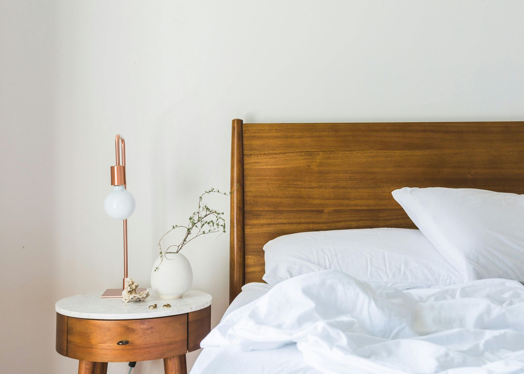 Bedroom scene with a wooden headboard, white bedding, and a nightstand with a lamp and vase.