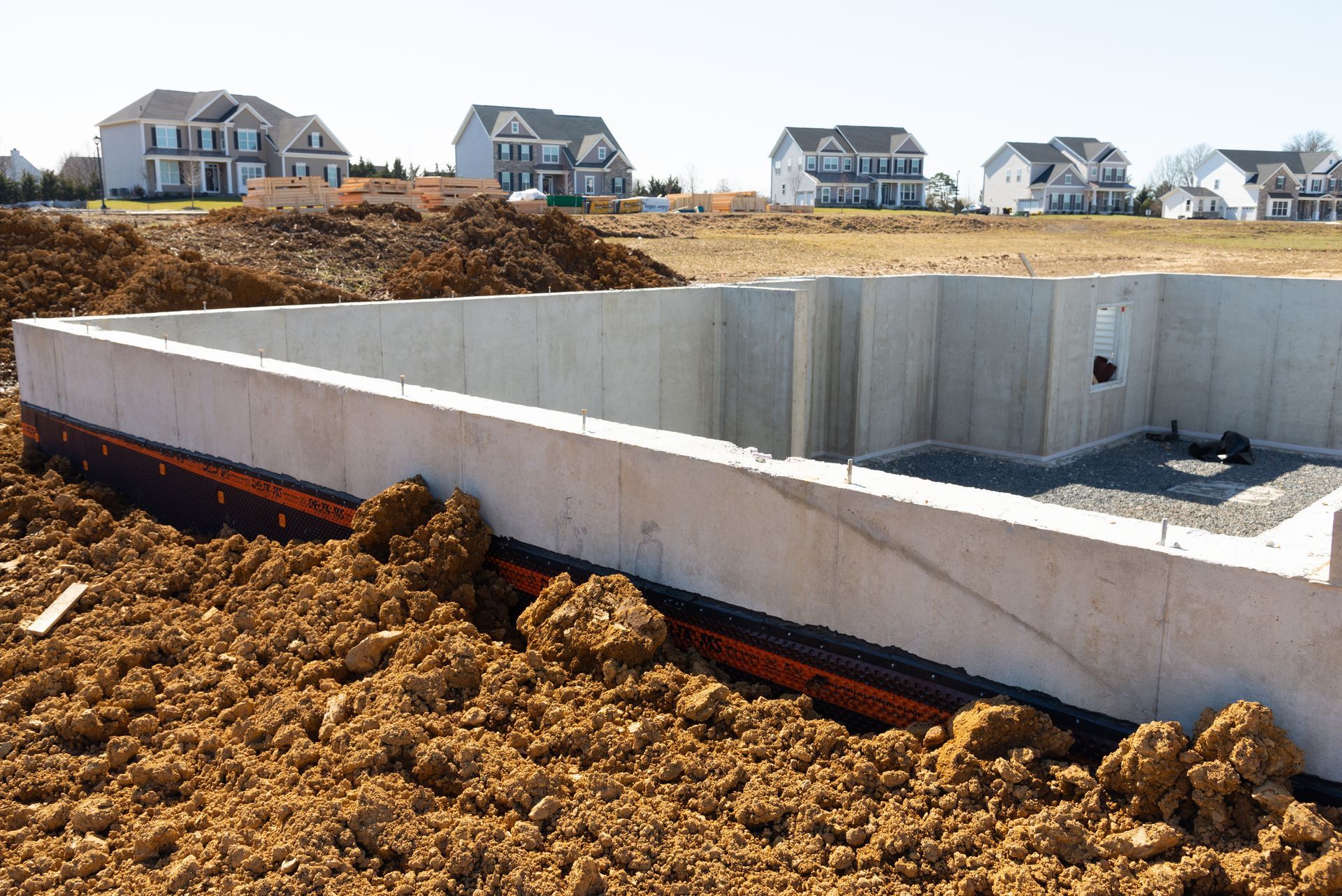 Murs de fondation en béton sur un chantier de construction, entourés de terre, avec des maisons en arrière-plan.