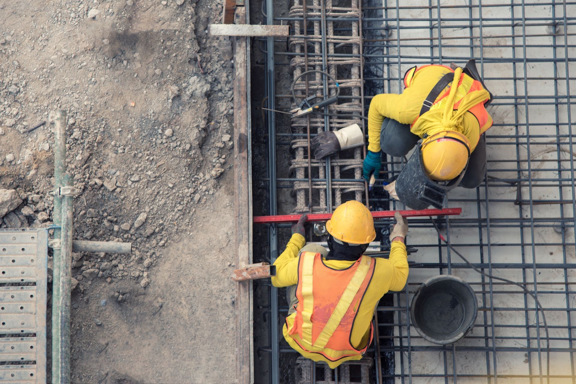 Deux ouvriers du bâtiment, vêtus de gilets jaunes et de casques de chantier, travaillent sur des barres d'armature, vue de dessus.