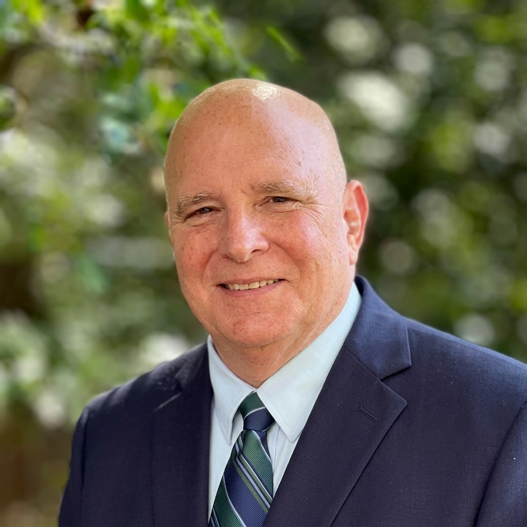 Headshot of a smiling bald man in a navy suit and striped tie against a blurred green background