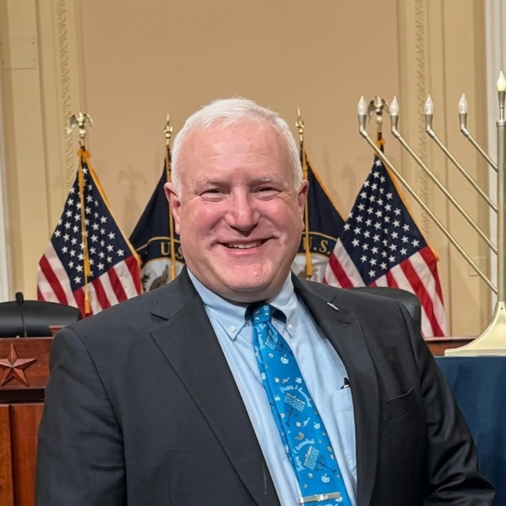 Smiling man in suit and blue tie standing before U.S. flags in a formal hall