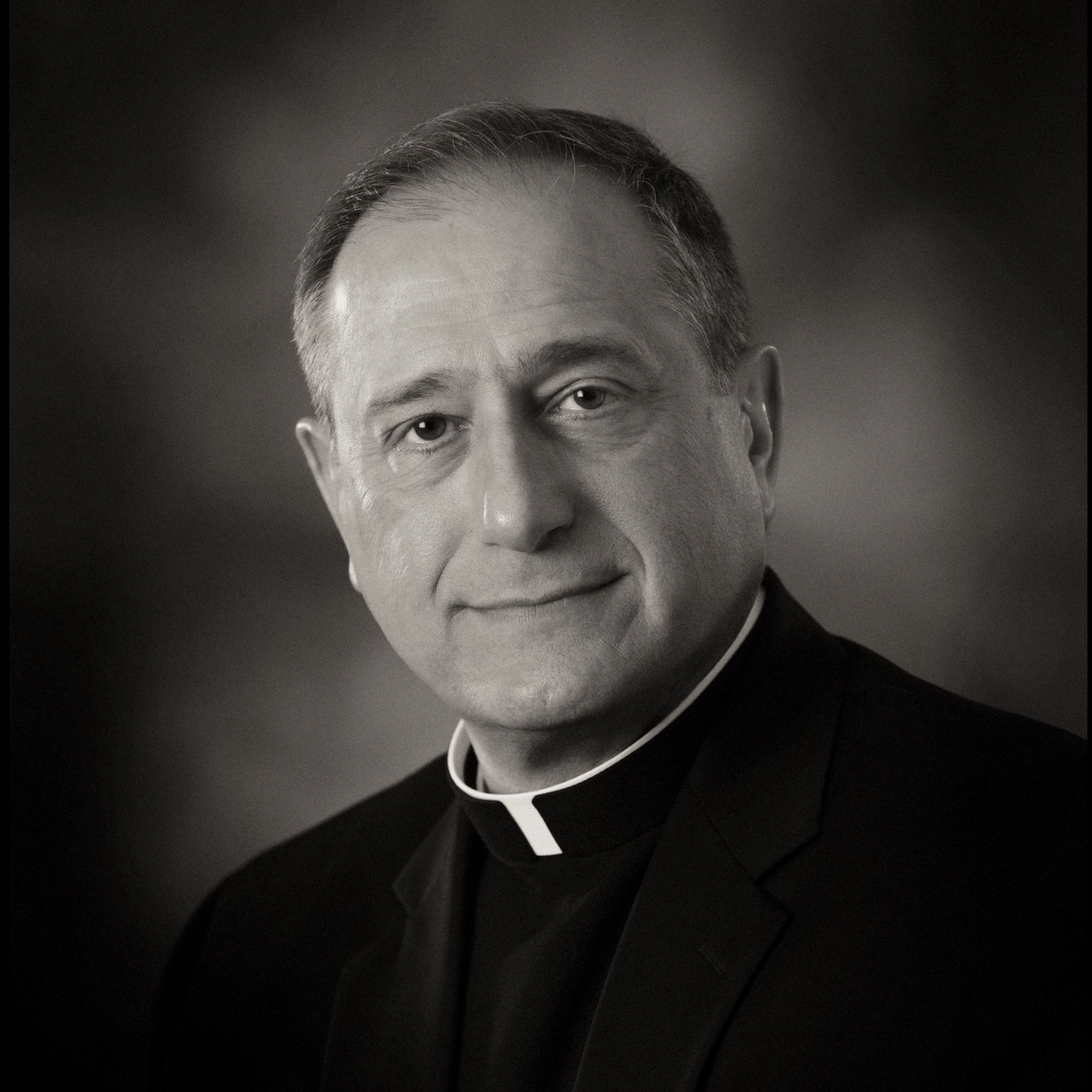 Black-and-white portrait of a smiling Catholic priest in clerical collar, against a dark background