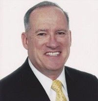 Portrait of a smiling man in a black suit, white shirt, and yellow tie against a light background