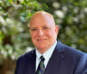 Smiling bald man in a dark suit and striped tie against a blurred green outdoor background
