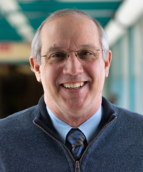 Smiling man wearing glasses, a blue sweater, and tie, standing in a blurred indoor hallway.