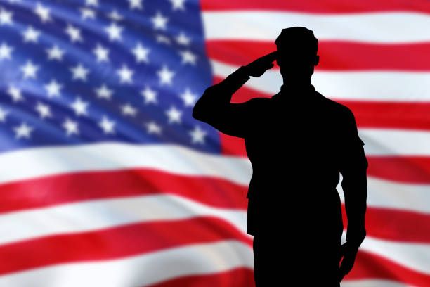 Volunteer at check-in table with Military Aid boxes and small flags, in front of a U.S. flag backdrop.
