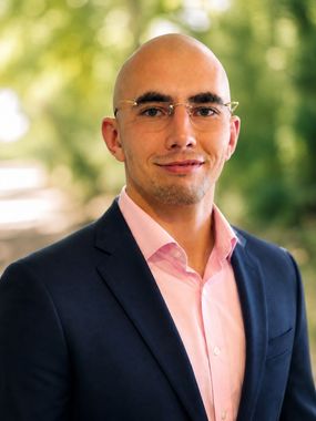 Portrait of a bald man in a navy blazer and pink shirt, standing outdoors with a blurred green background