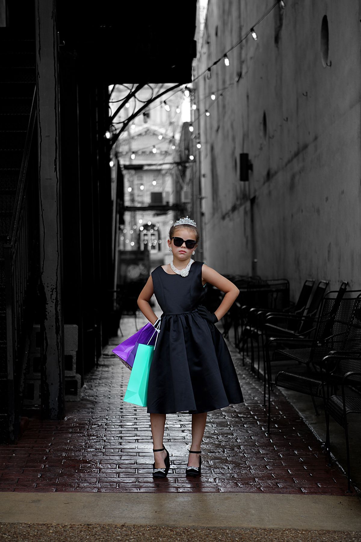 A little girl in a black dress is standing in an alleyway holding shopping bags.