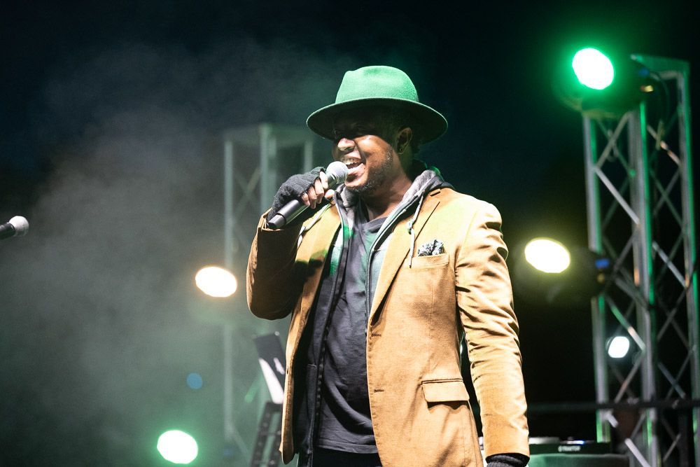 DJ & MC in a tan coat and green hat performing on stage with a microphone under bright lights at Colorado State University