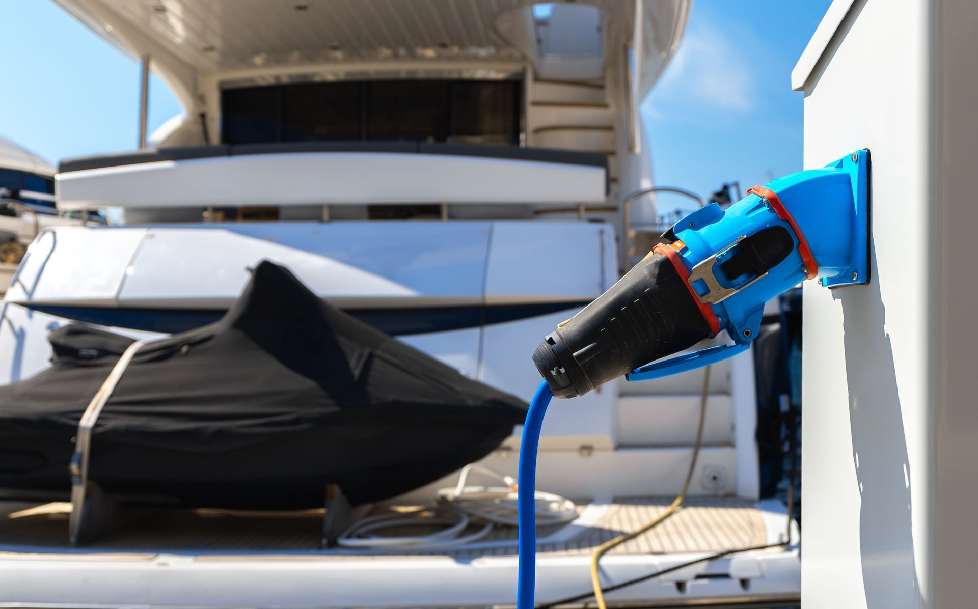 Blue power cable plugged into a white dockside electrical outlet, with a boat in the background.