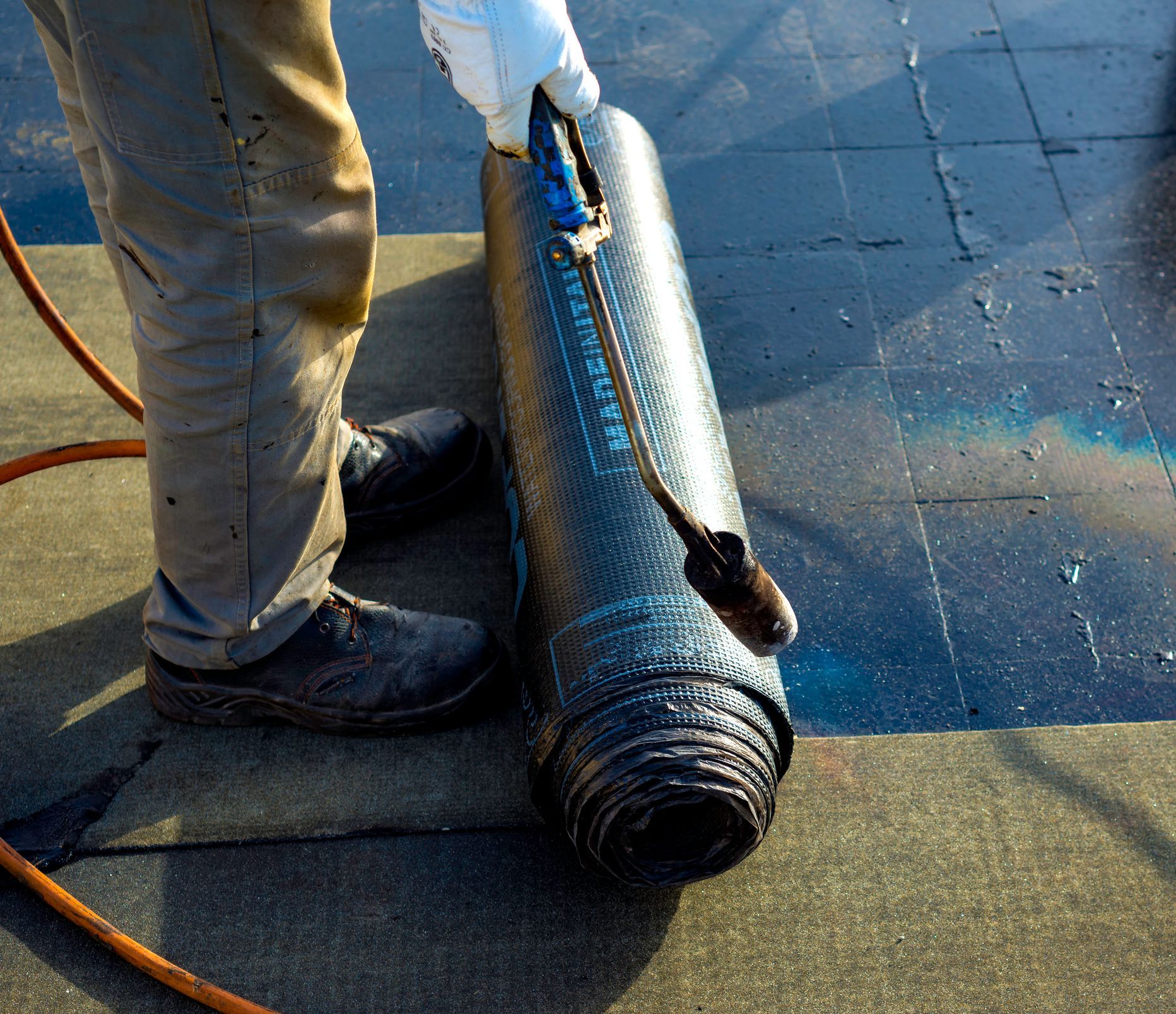 Un operaio utilizza una torcia a propano per installare un rotolo di materiale bituminoso nero per tetti su una superficie piana.