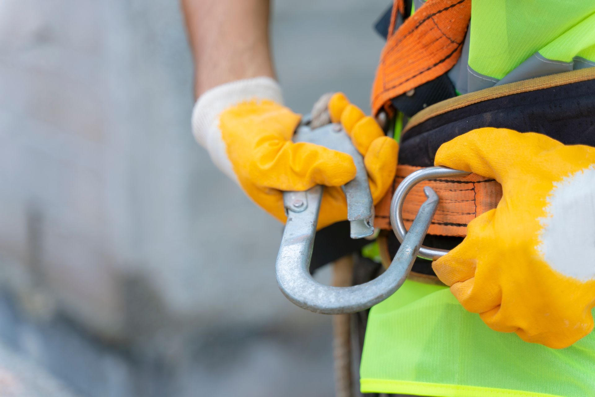 Un operaio con giubbotto ad alta visibilità e guanti gialli fissa un gancio metallico di sicurezza a una cinghia.