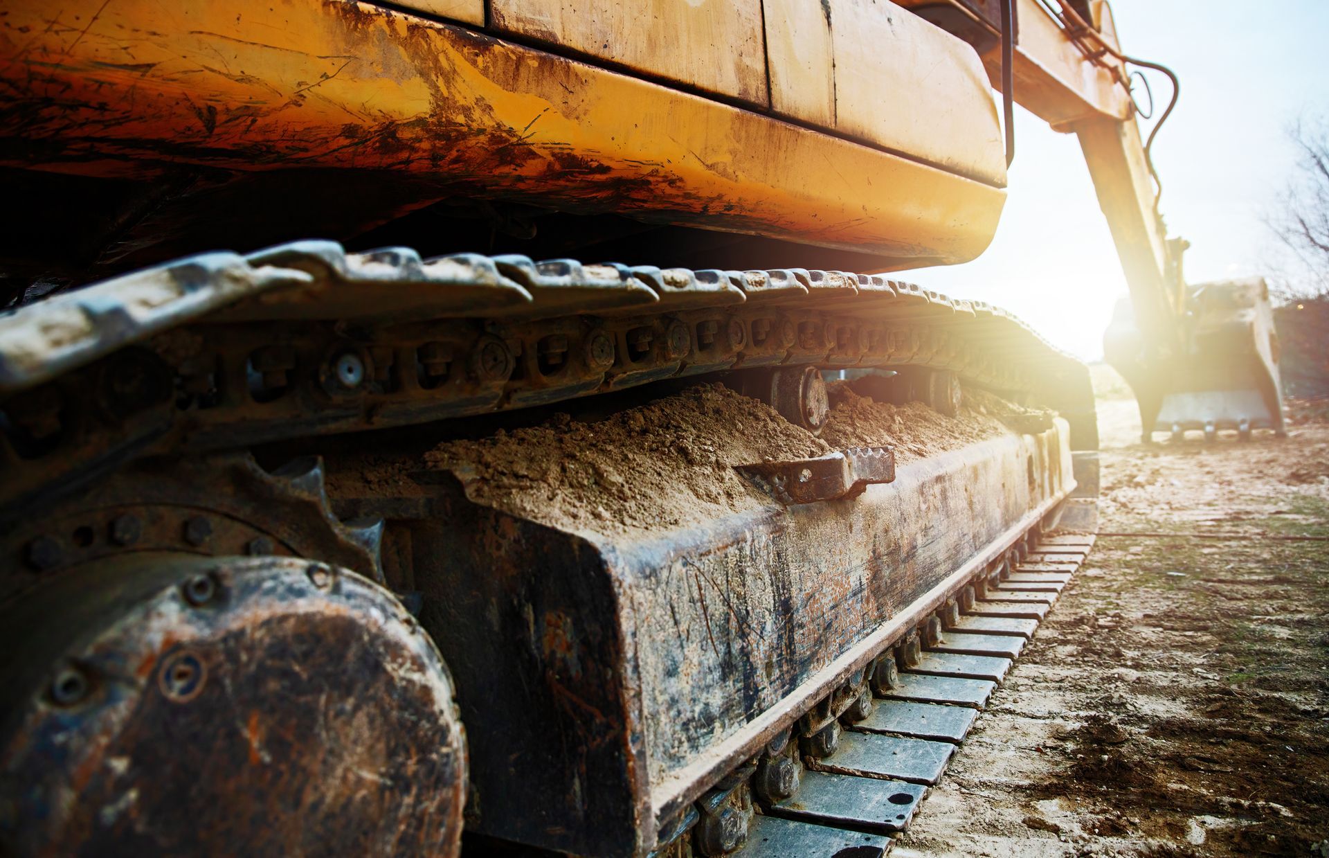 Primo piano dei cingoli arancioni di un escavatore appoggiati sul terreno in un cantiere, con la luce del sole che si riflette sullo sfondo.