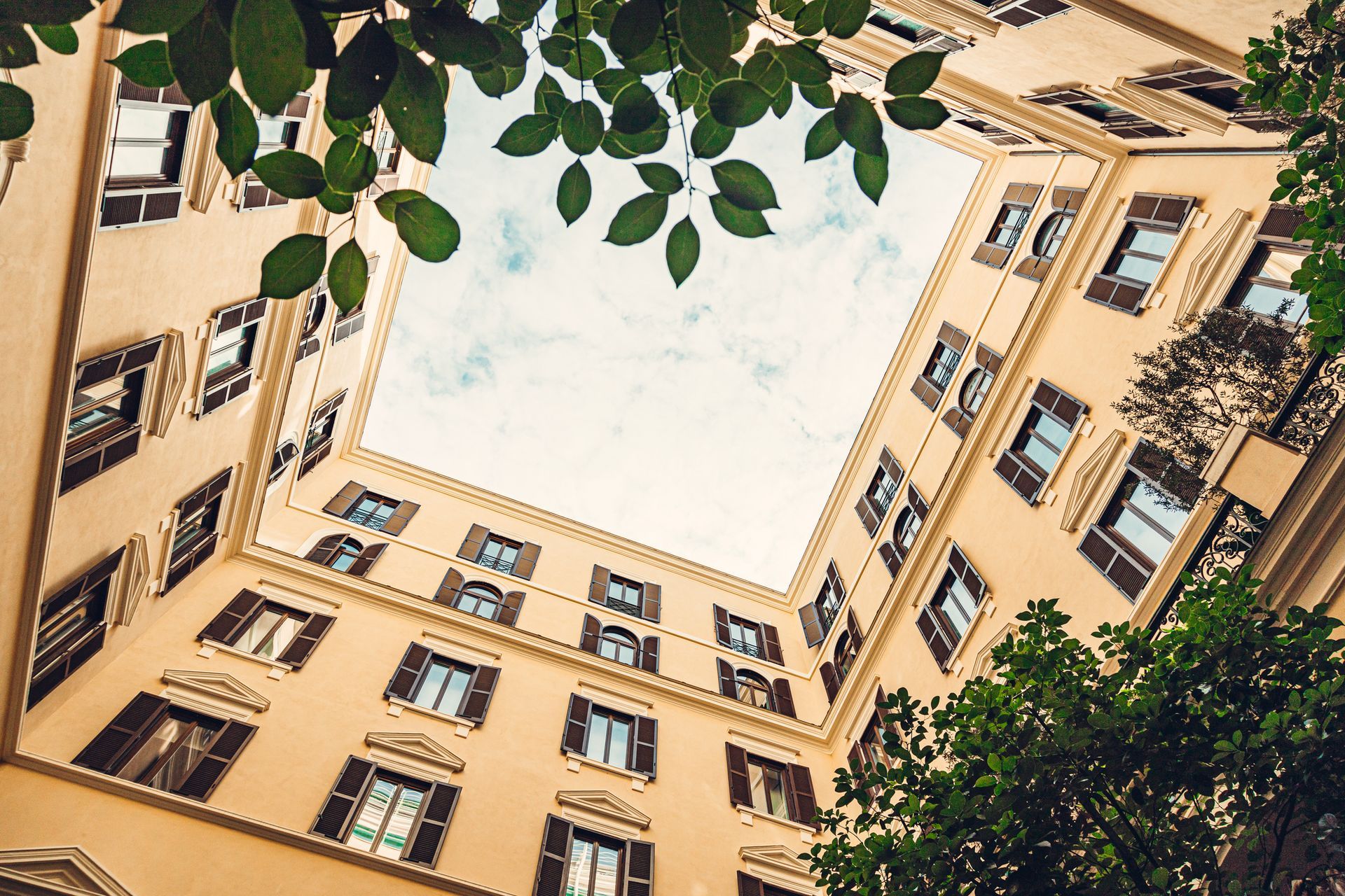 Una veduta dal basso del cortile di un edificio beige, incorniciato da foglie verdi, con lo sguardo rivolto verso un cielo luminoso e nuvoloso.