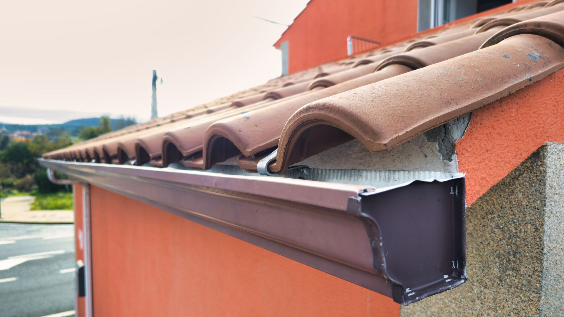 Primo piano di una grondaia metallica rettangolare di colore marrone, fissata al bordo del tetto di una casa con tegole in terracotta.