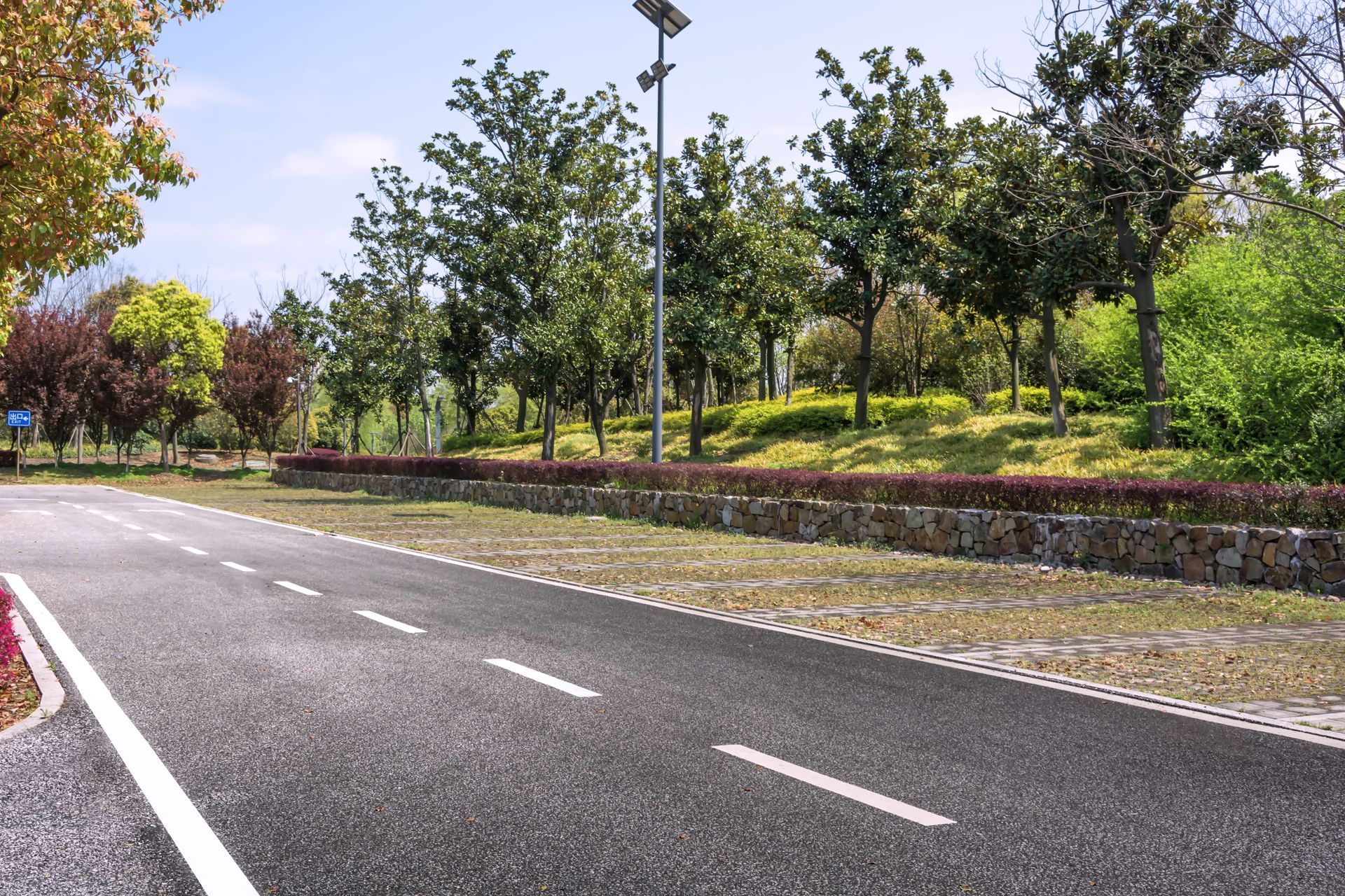 A paved road with white dashed lines curves alongside a stone retaining wall, lush green trees, and shrubs.