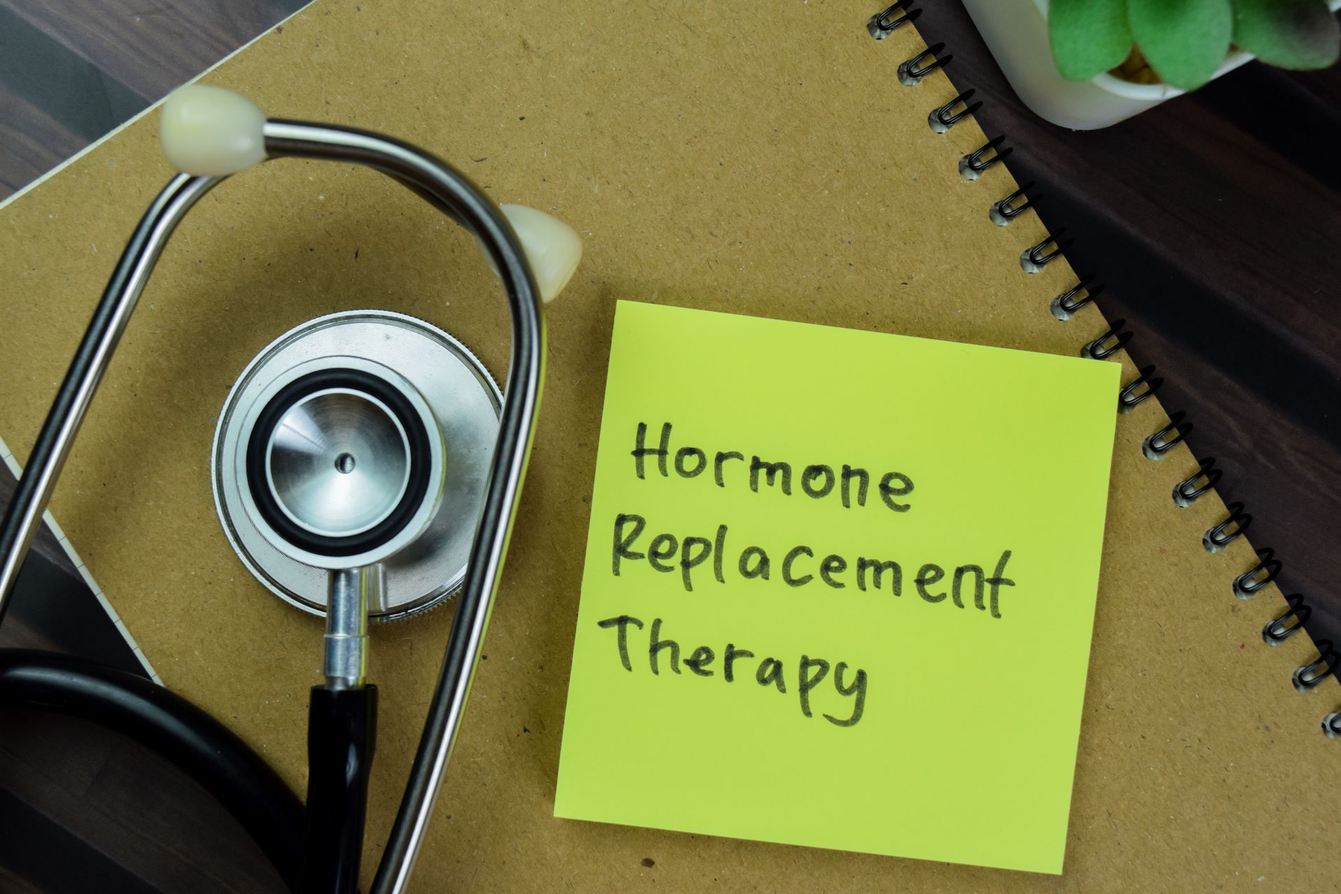 A stethoscope lies next to a lime-green sticky note on a brown notebook, with the text Hormone Replacement Therapy. A stethoscope lies next to a lime-green sticky note on a brown notebook, with the text Hormone Replacement Therapy.