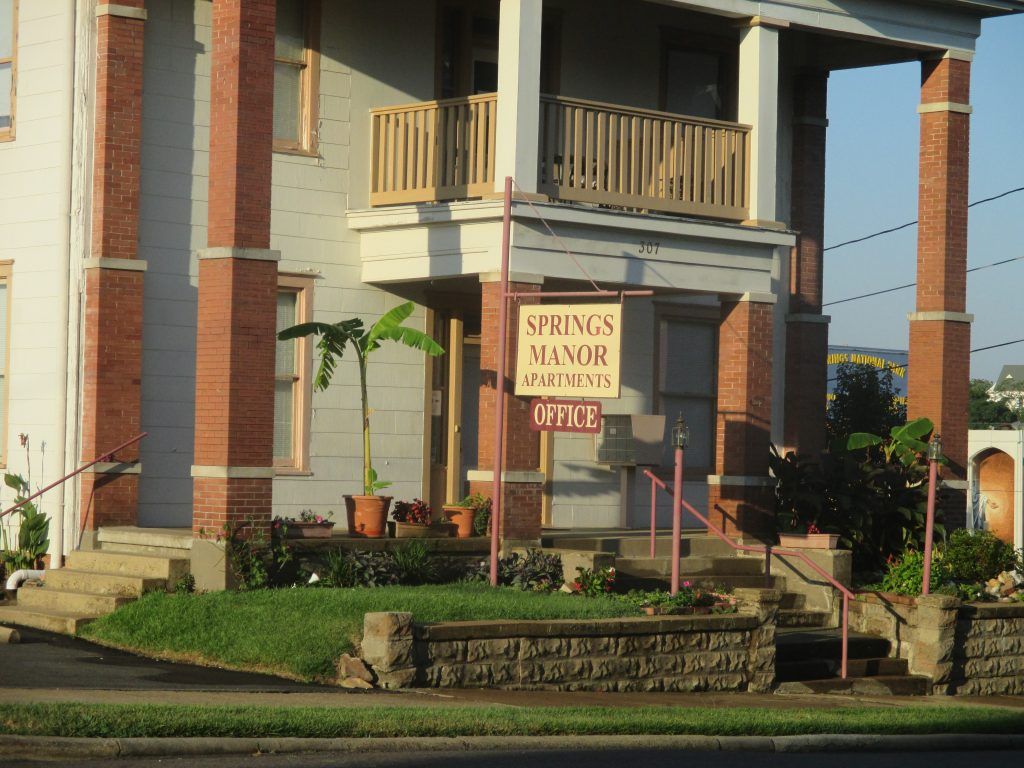 Springs Manor Apartments building with brick columns, sign, and small garden.
