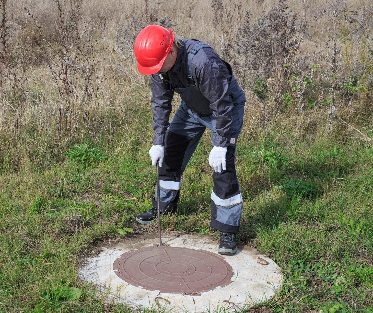 Person in red hardhat and overalls checks a manhole in a grassy field.