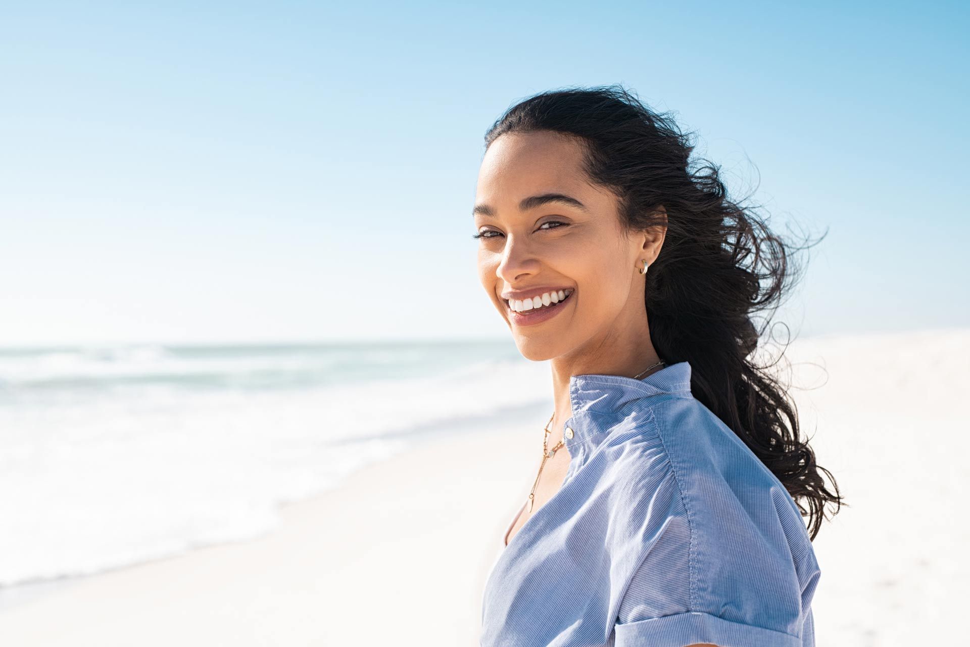Woman smiling on a beach, wearing a blue striped shirt, with the ocean and blue sky in the background.