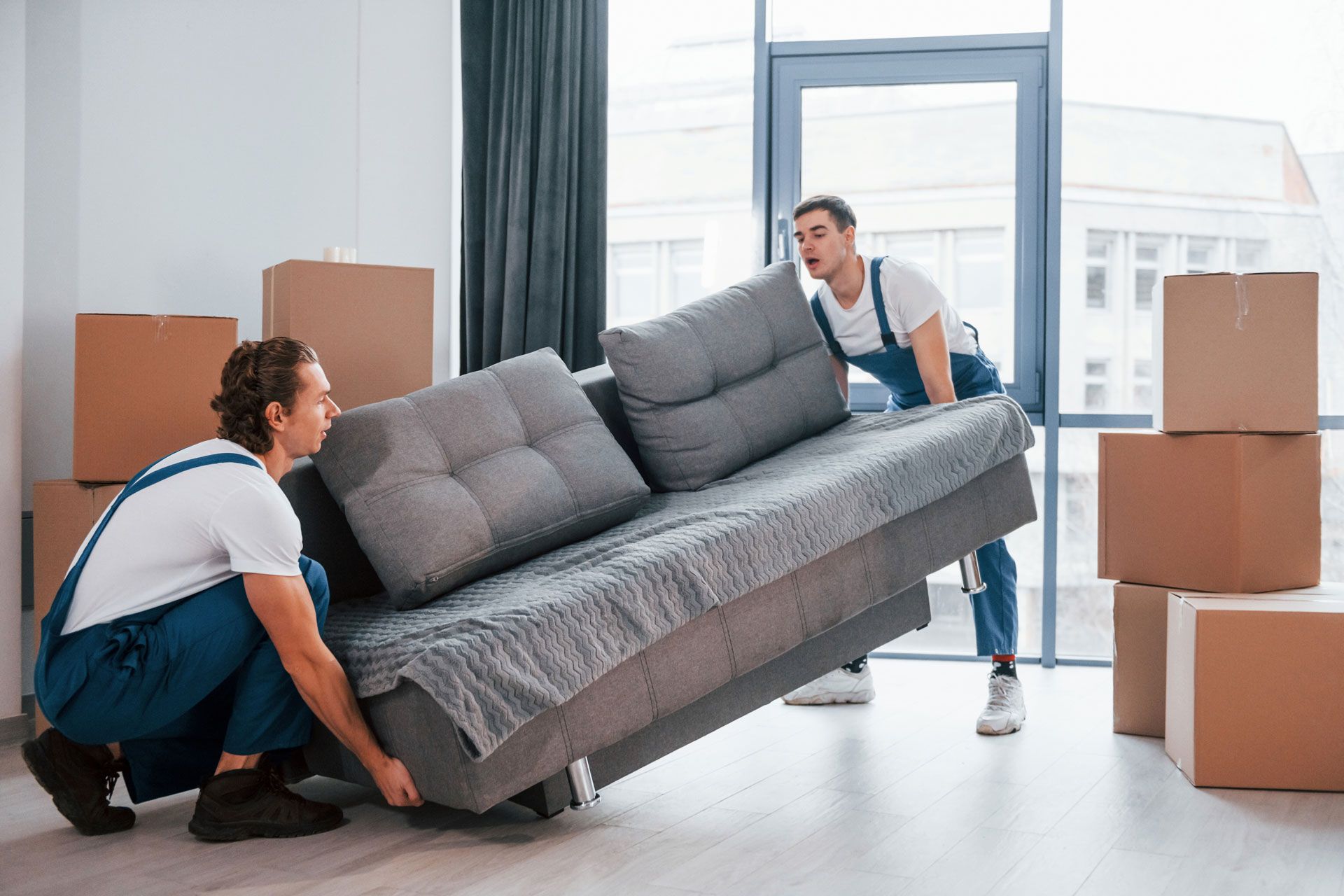 Two movers in blue overalls lift a gray sofa in a room with boxes.