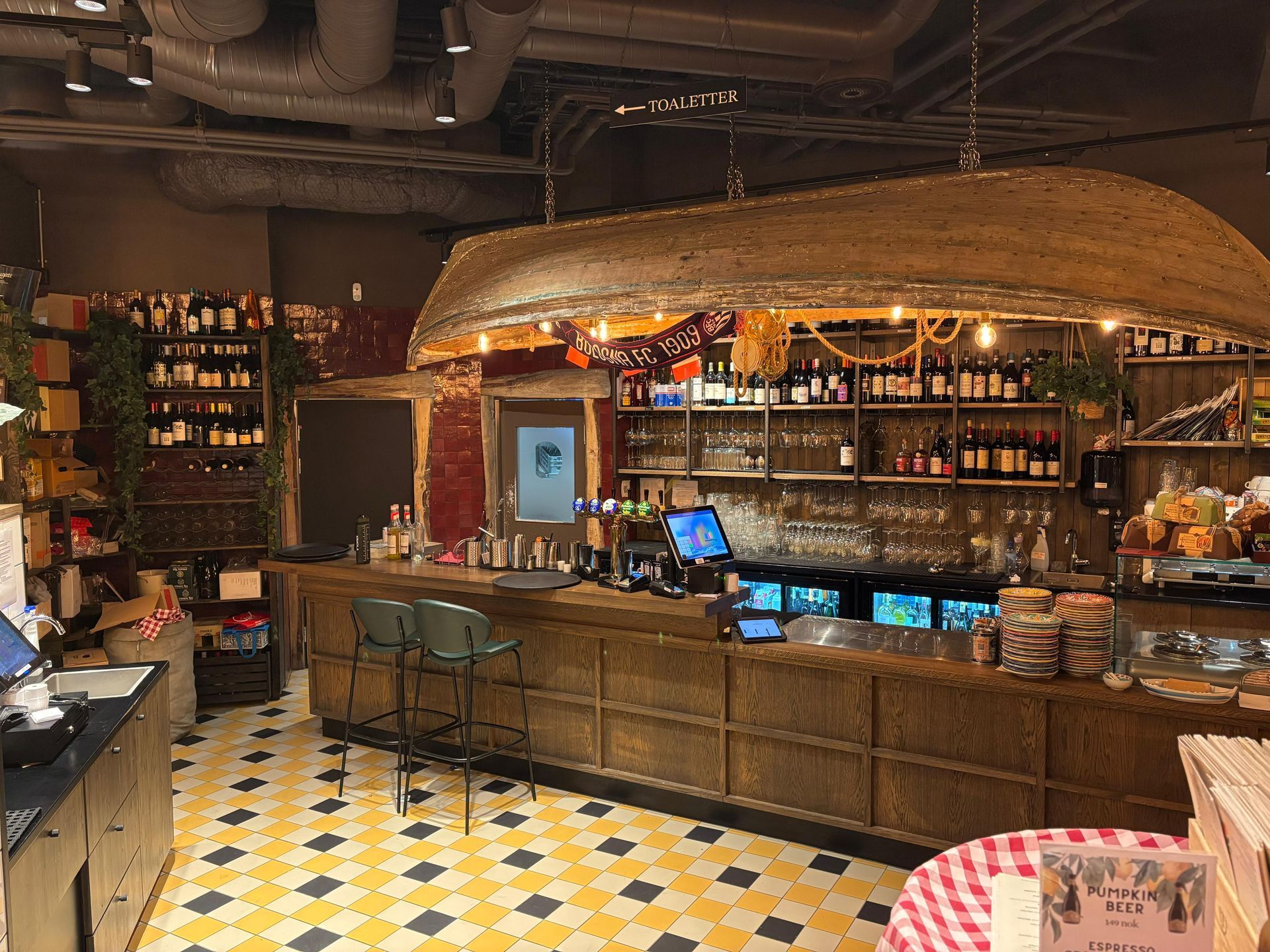 Bar interior with wooden counter, canoe-shaped light fixture, bottles on shelves, checkered floor.