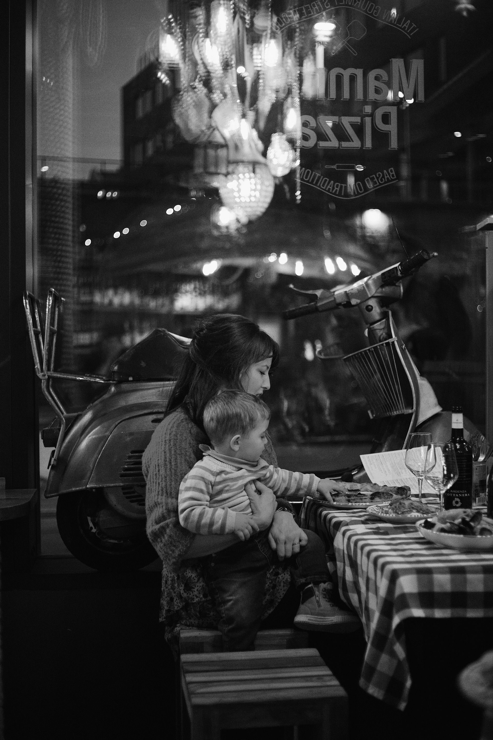 Woman and toddler at a restaurant table with scooter visible through the window, black and white.