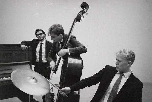 Black and white photo of a jazz trio: pianist, bassist, and drummer in suits, playing instruments.